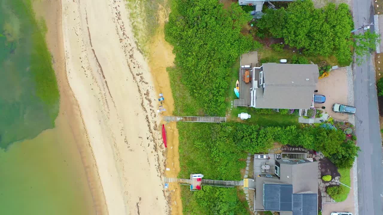 Scenic top-down drone view of Cape Cod shoreline homes, sandy beach, ocean waves, and surrounding greenery, showing neighborhood roads and walkways to the waterfront