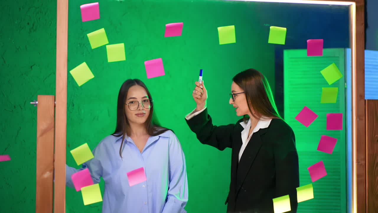 Caucasian women with long brunette hair wearing glasses stand at glass board. Positive ladies take stickers off the glass and throw them away.