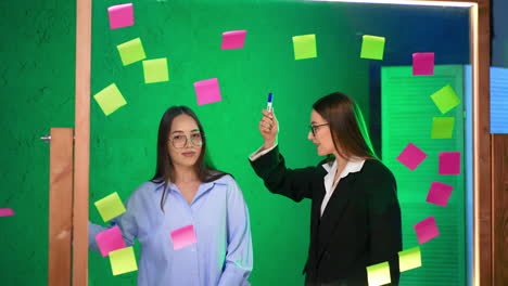 caucasian women with long brunette hair wearing glasses stand at glass board. positive ladies take stickers off the glass and throw them away.
