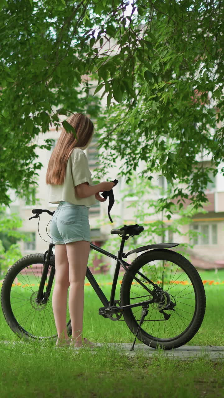 여성은 포장된 길에 주차된 검은색 자전거에 접근하고, 좌석과 손에 장갑을 착용합니다. 배경은 높은 나무와 주거 건물로 둘러싸인 멀리 달리는 아이를 보여줍니다.