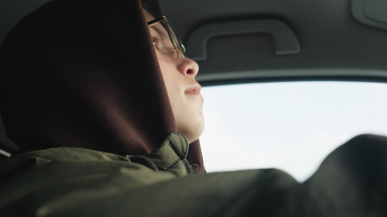 close up of boy in hoodie wearing glasses staring thoughtfully through car window with soft expression and blurred background of high rise buildings
