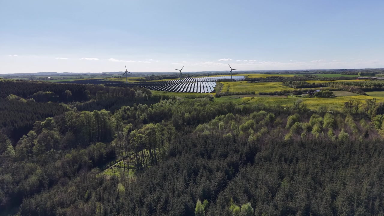 A tracking drone shot of a combined solar and wind power plant integrated into yellow rapeseed fields in the rural Danish countryside near Hammel.