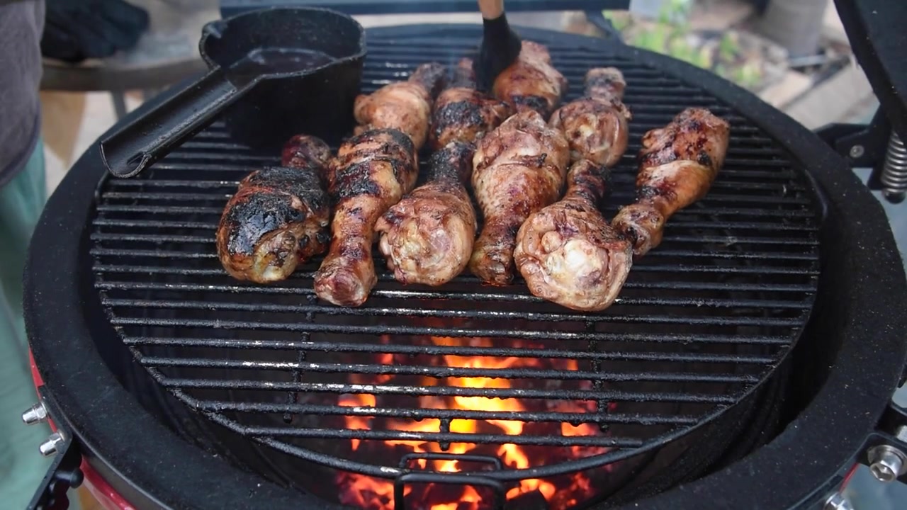 Fusi di pollo che grigliano su carbonella ardente in un affumicatore barbecue stile kamado. Uno chef spennella una marinata saporita sulla carne durante la cottura