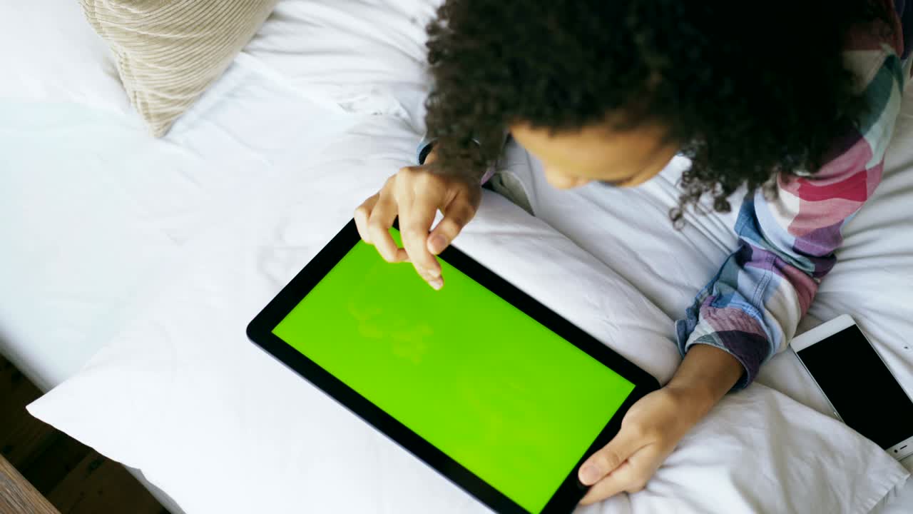 vista superior de una mujer de raza mixta rizada acostada en la cama en casa usando una tableta electrónica con pantalla verde