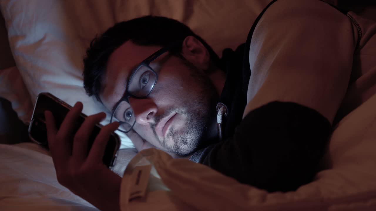 hombre acostado en la cama con un teléfono inteligente, la pantalla ilumina la cara en un dormitorio oscuro, desplazándose, redes sociales, enviando mensajes de texto a amigos en la noche antes de dormir