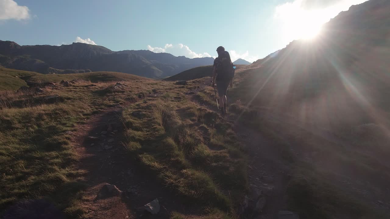 vista trasera de un joven excursionista con mochila y bastón de trekking caminando hacia el sol en los pirineos españoles durante la puesta de sol