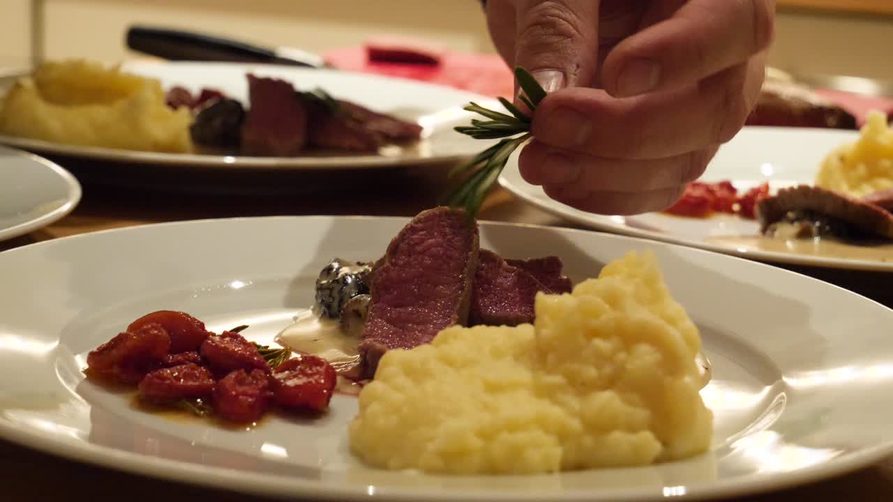un modelo de mano masculino blanco está poniendo romero en la carne roja de cordero junto al puré de papas y tomates en cámara lenta