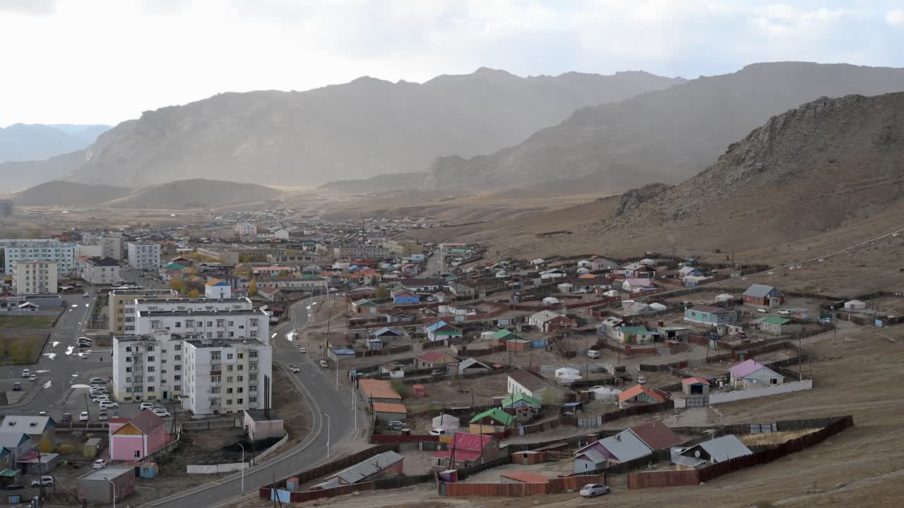 An atmospheric, high-angle shot of Uliastai, Mongolia, as wind and dust sweep through the mountain valley. Sunlight breaks through the haze, illuminating the city's diverse architecture
