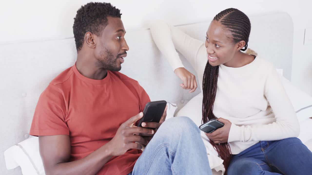feliz pareja afroamericana sentada en la cama hablando y usando teléfonos inteligentes, en cámara lenta
