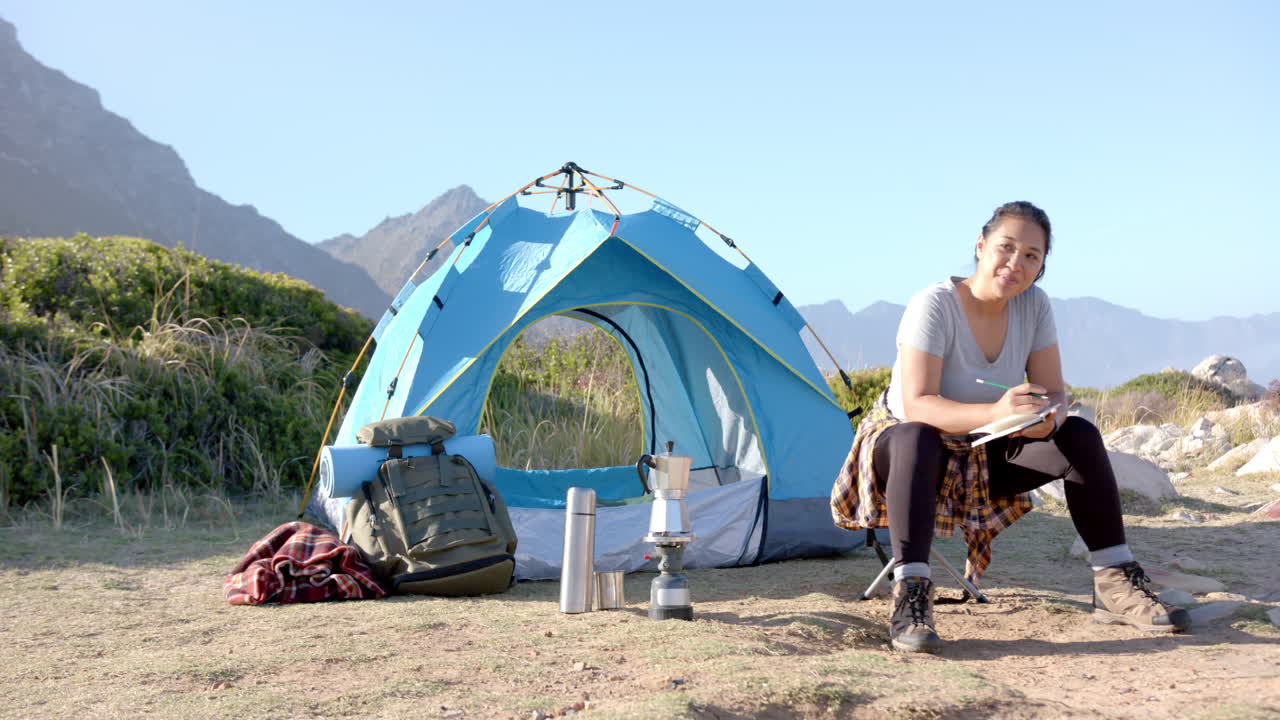 Camping in mountains, woman sitting by tent and writing in notebook