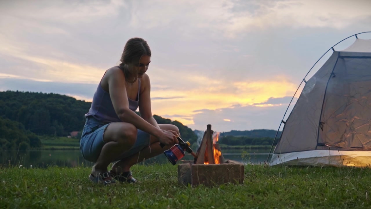 Kvinde der sidder på hug i græsset og bruger en butangasbrænder til at tænde et lejrbål nær telt og rolig sø ved solnedgang, der fanger udendørs campingaktivitet og fredelig naturlig atmosfære i Savci, Slovenien.