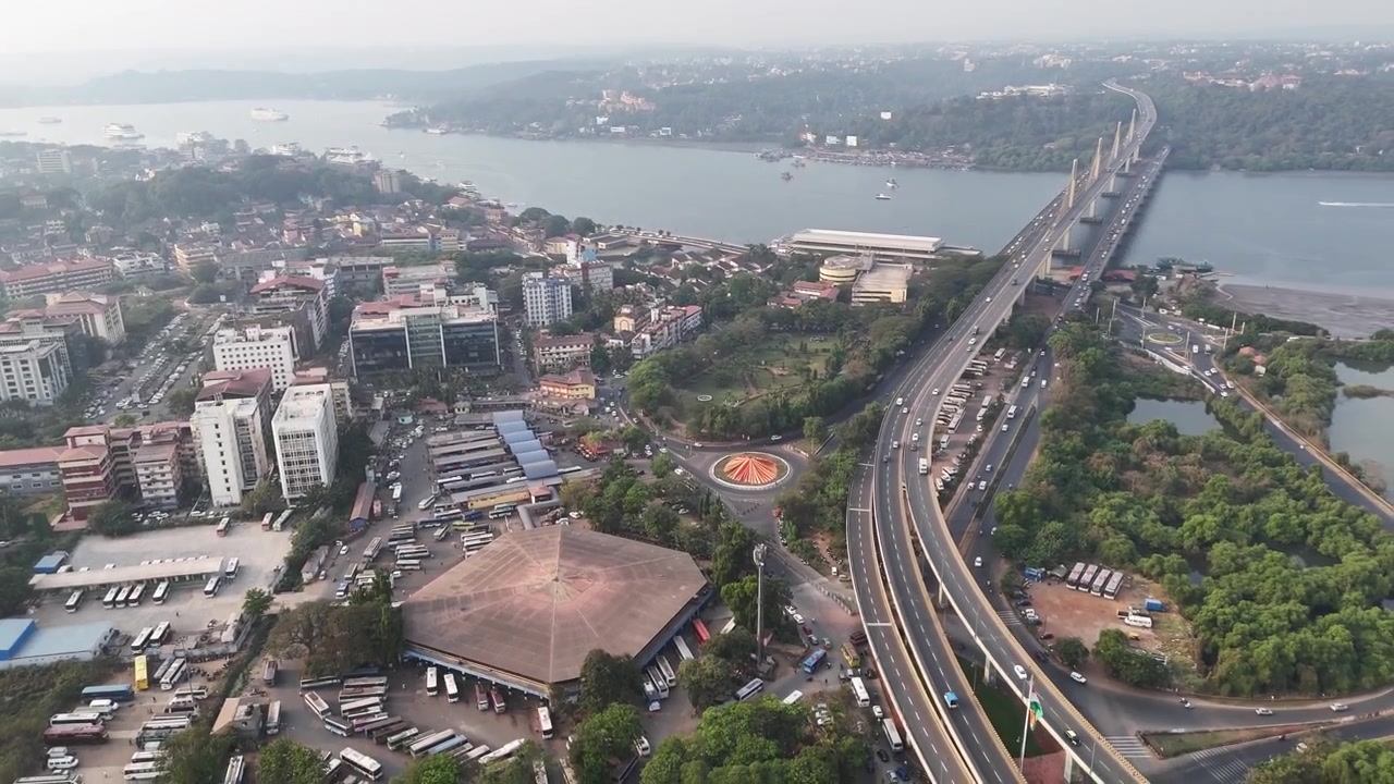 Ampia ripresa aerea cinematografica di un paesaggio urbano durante l'inverno che cattura autostrade curve e incroci trafficati a Panaji KTC Bus Stand, Goa, che conducono verso Atal Setu, con viste sul fiume e vegetazione che aggiungono profondità
