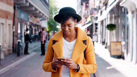 Mujer-Negra-Milenaria-Con-Un-Abrigo-Amarillo-Y-Un-Sombrero-Homburg-Usando-Su-Teléfono-Inteligente-Parada-En-Una-Calle-Y-Saliendo-De-La-Toma,-De-Cerca