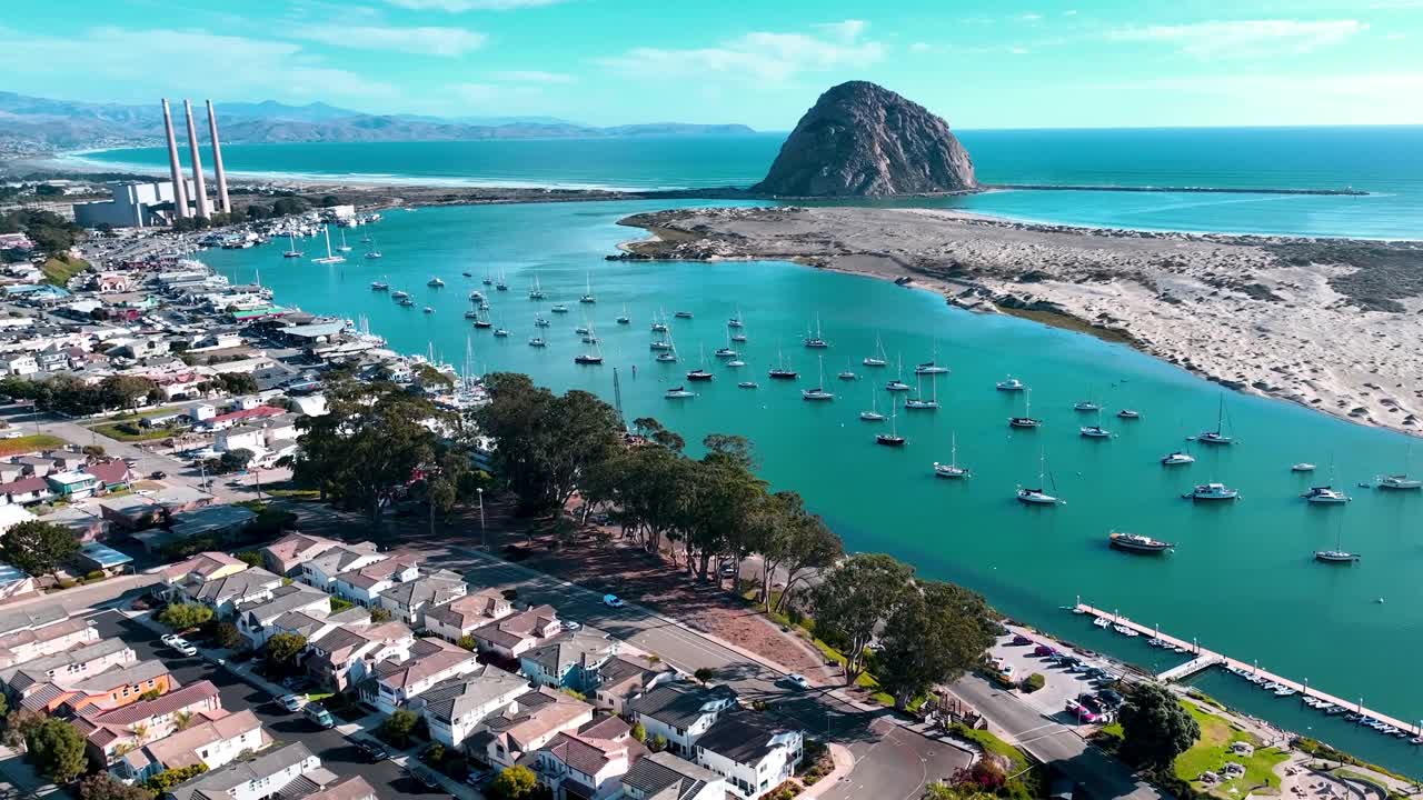 Kaliforniya'nın Merkez Sahili'ndeki Morro Bay Limanı, Morro Kayası, marina tekneleri, kıyı evleri ve eski enerji santralinin berrak mavi gökyüzü altında çekilmiş drone görüntüsü.