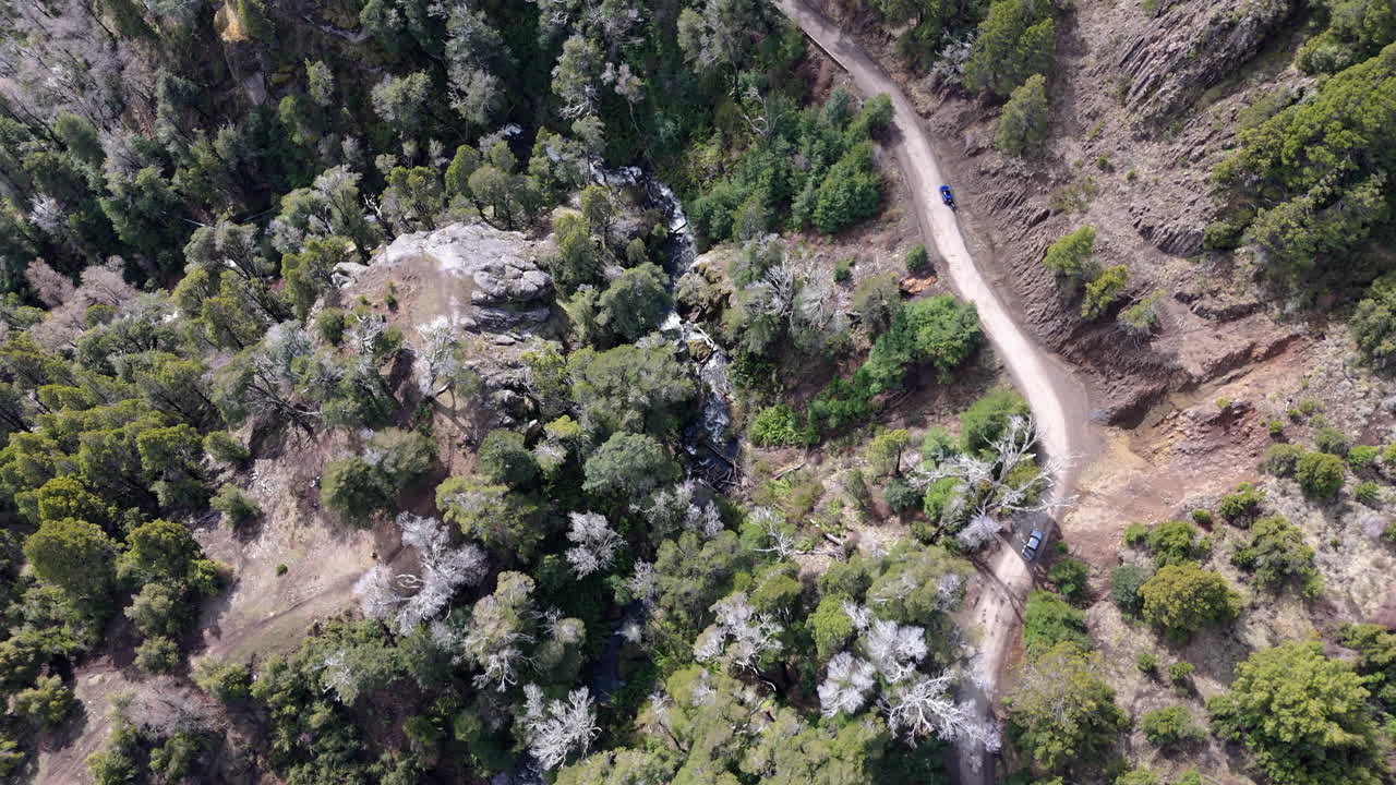 Aerial view of a muddy road winding through a dense patagonian forest, a small waterfall cascading nearby, San Martin de los Andes, Neuquen, Argentina, overehad drone shot