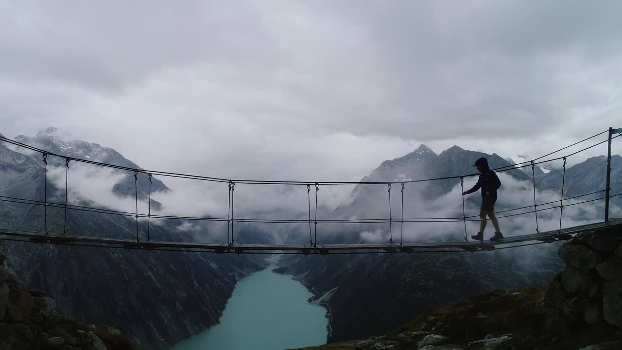 Hiker walks hanging bridge above turquoise lake and misty peaks in moody Austria