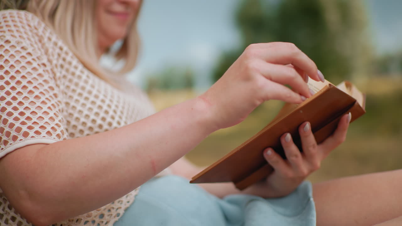 Close-up side view of woman reading old book outdoors, flipping pages with delicate hands, soft blur on blue shorts and rustic background