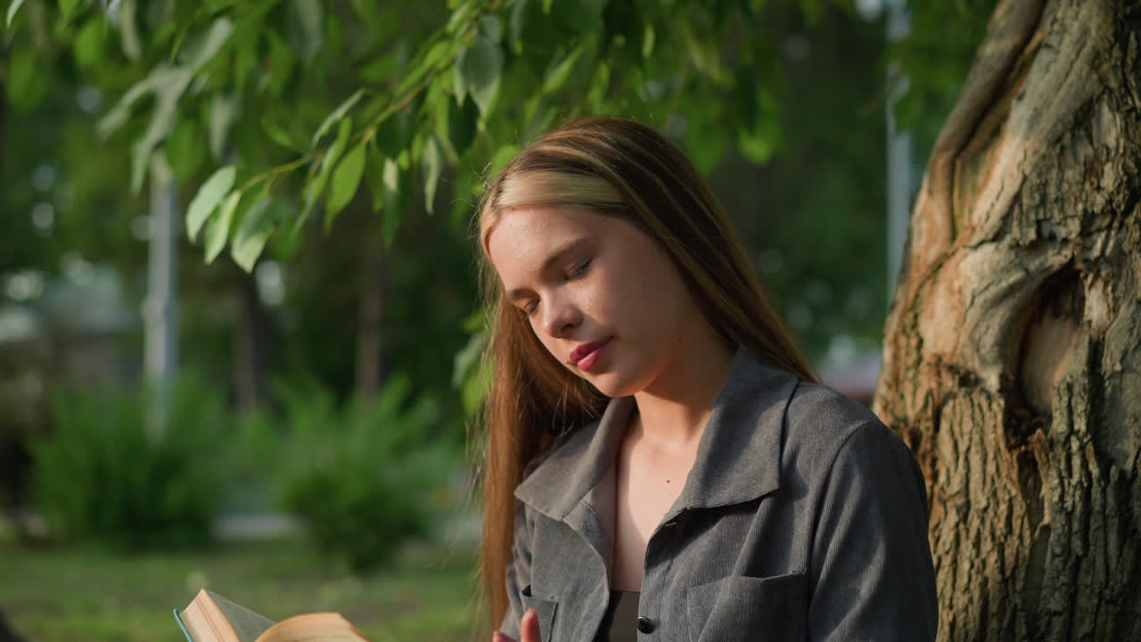 야외에 앉아서 책을 손에 고, 머리를 약간 오른쪽으로 기울여, 사려 깊게 보이는데, 배경에는 초록색이 있고, 빛이 부드럽게 그녀의 얼굴을 비추고 있다.