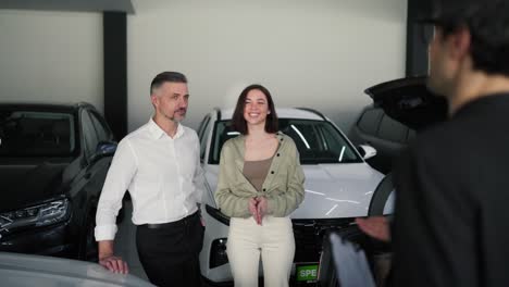 sobre su hombro un hombre confiado de mediana edad con una barba gris y su esposa morena en una sala de exhibición de automóviles están hablando con un asistente en traje sobre modelos de automóviles modernos y escuchando automóviles en una salón de exhibiciones de automóviles