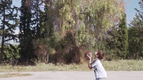 niño caucásico juega baloncesto en la cancha de baloncesto del callejón, árboles altos en el fondo, cámara lenta