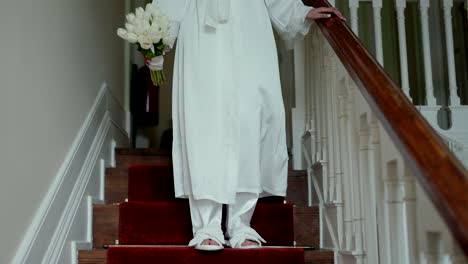 bride descends carpeted stairs in robe with wedding bouquet in hand