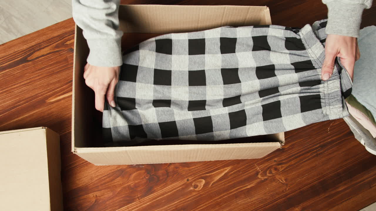 Hands Packing Folded Checkered Clothes into Cardboard Box