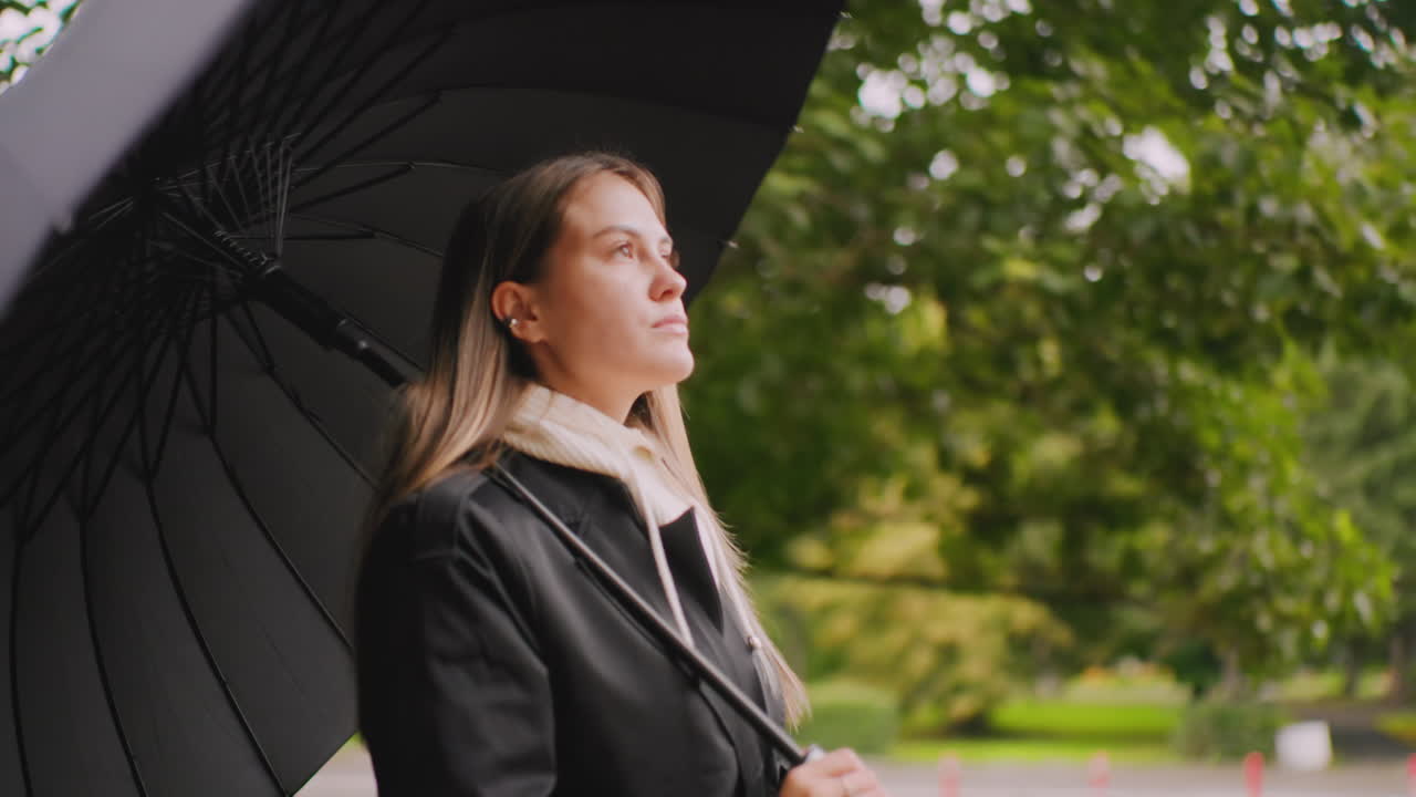 Young woman in black coat holding umbrella standing outdoors on rainy day, looking thoughtful with calm expression, surrounded by green trees in park, peaceful autumn atmosphere with overcast sky