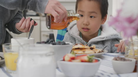 Lindo-Niño-Asiático-Comiendo-Gofres-Frescos-Para-El-Desayuno-Disfrutando-De-Una-Deliciosa-Comida-Casera-Con-La-Familia-En-La-Cocina-De-Casa-4k