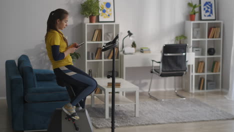 Una-Joven-Deportista-Descansa-Durante-El-Entrenamiento-Con-Una-Bicicleta-De-Ejercicio-Usando-Redes-Sociales-En-Un-Teléfono-Inteligente,-Equipo-De-Fitness-En-Casa,-Adicción-A-Internet