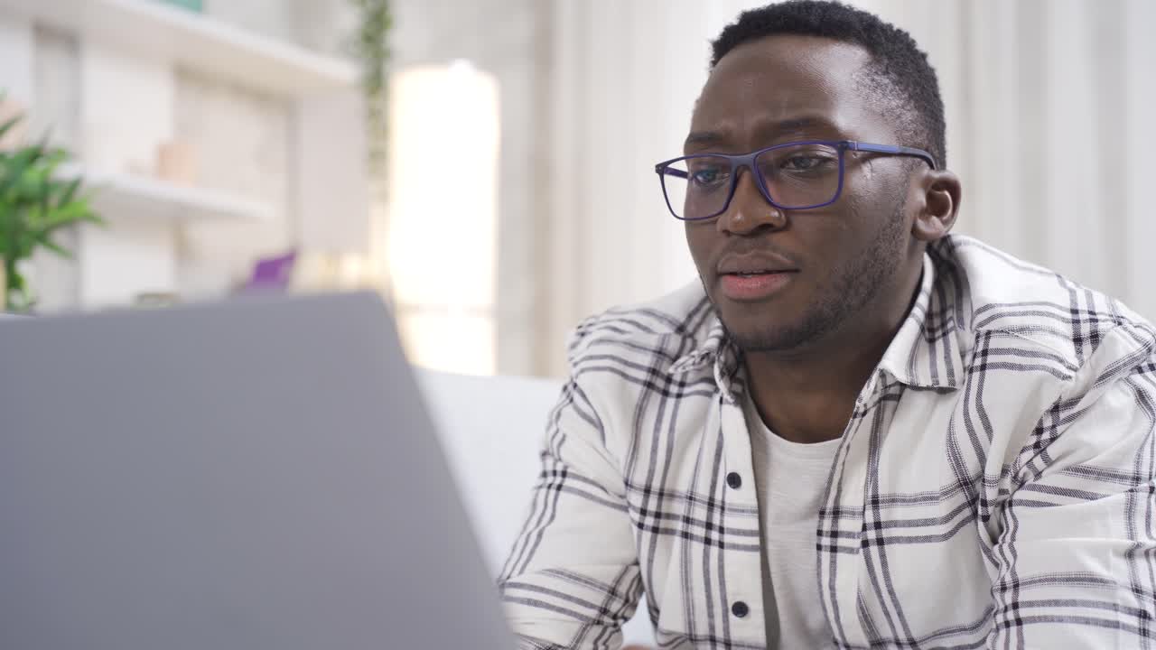 Handsome happy african american young man working at home on sofa looking at laptop