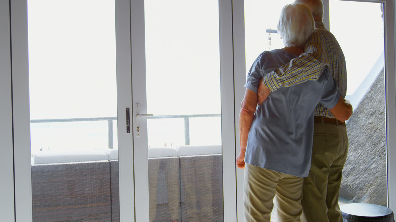 vista trasera de una pareja de ancianos caucásicos mirando por la ventana en una casa cómoda 4k