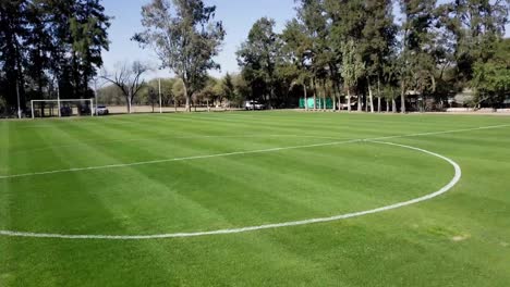 Drone-flying-low-over-an-empty-soccer-field-with-green-grass-and-trees-in-the-background