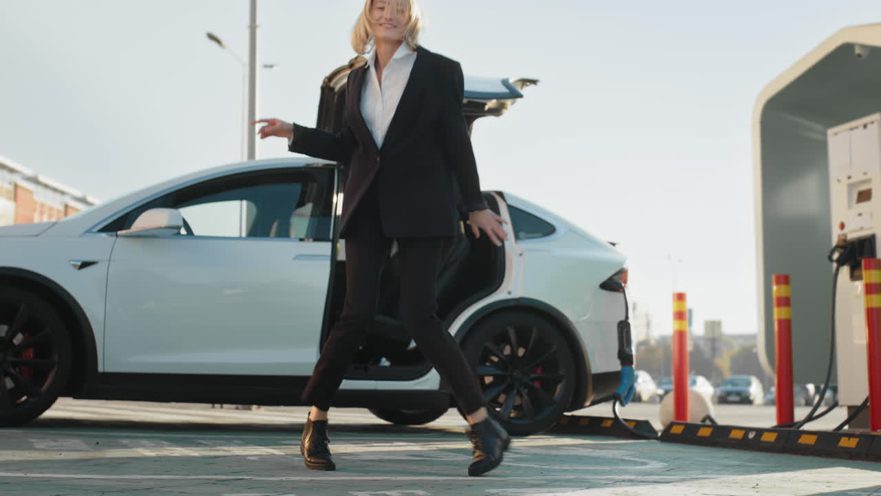 una mujer con un traje de negocios bailando felizmente mientras sale de su coche eléctrico blanco en una estación de carga.