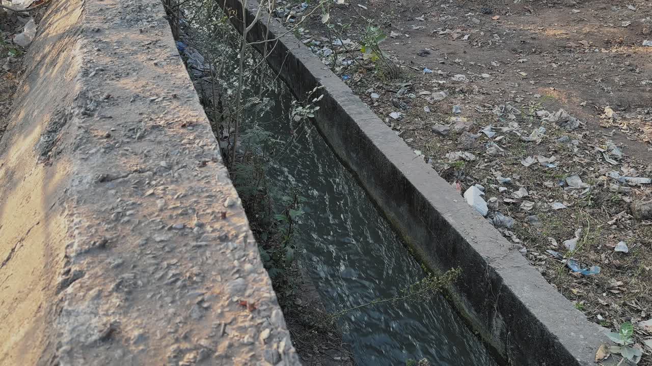 Static close shot of greywater flowing through a narrow concrete drain in India, murky stream bordered by rough walls, scattered litter, weeds, and dry soil along the edge