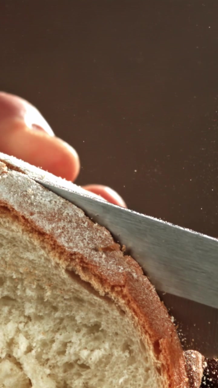 Close-up of a Serrated Knife Slicing Fresh Bread with Crumbs Flying in Slow Motion