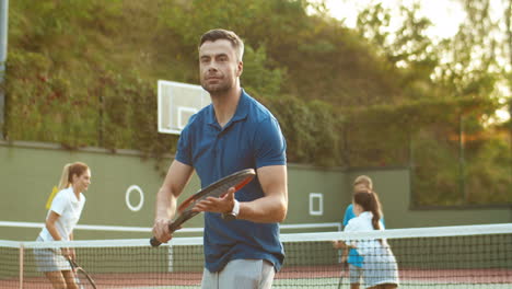 hombre guapo entrenando con raqueta mientras su familia juega al tenis en el fondo