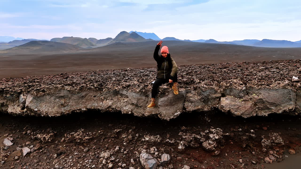 Woman sits on the rock edge waving her hand. Drone distances from tourist resting among the cosmic scenery of Iceland.