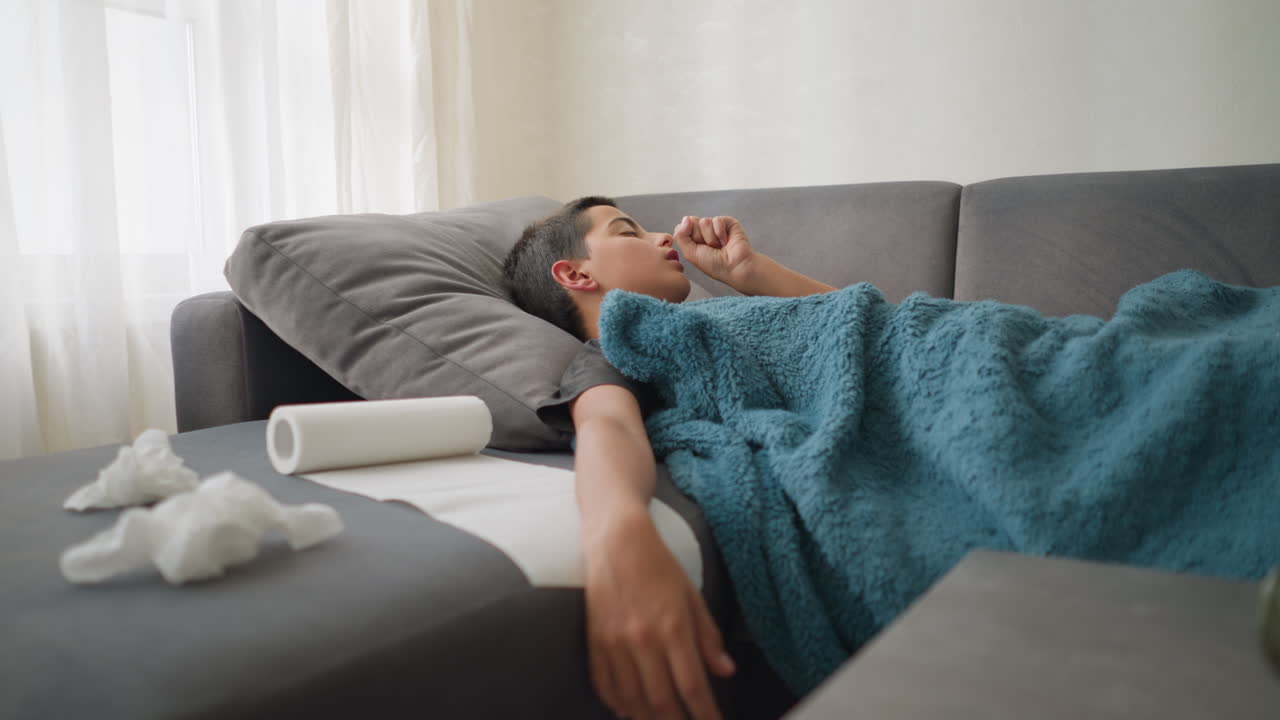 Sick child resting on couch with used tissue, thermometer, and glass cup on table, boy touches forehead with hand while covered in blanket, feeling unwell and exhausted in living room