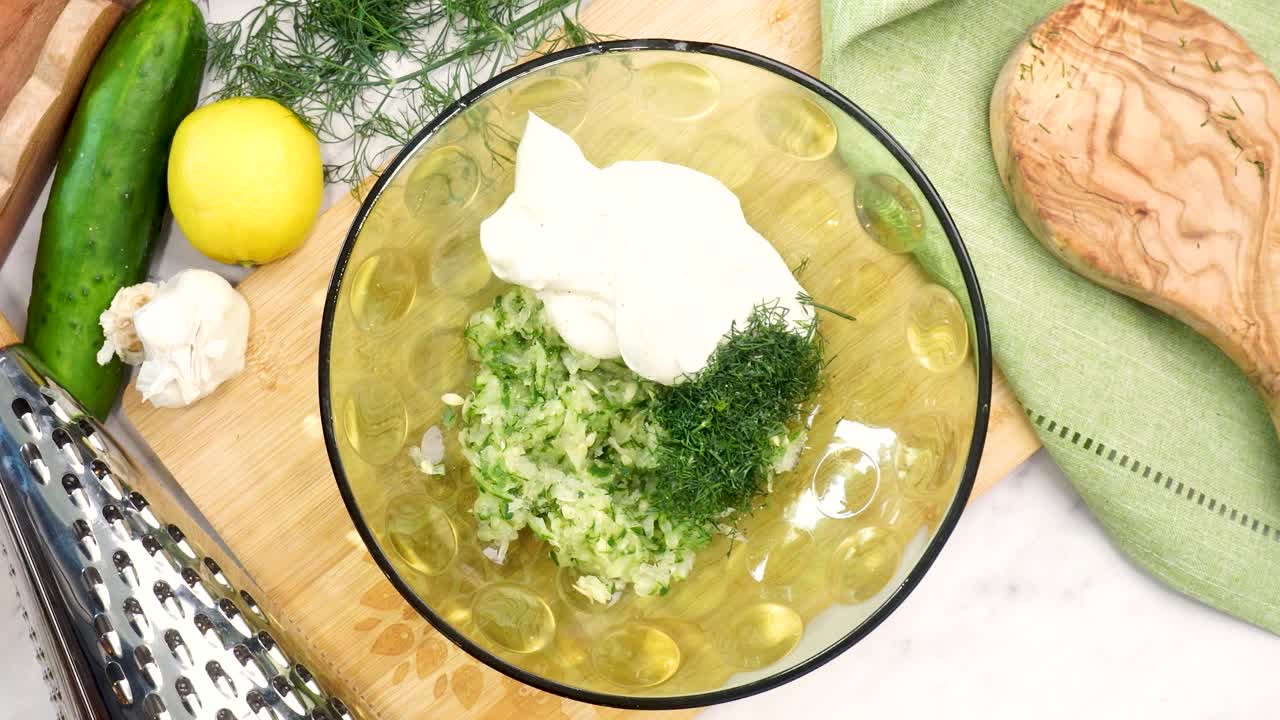 Making Tzatziki Greek or Mediterranean sauce or dip for a party. Dip or sauce in a green bowl on a wooden cutting board with dill, green napkin and a grater. Grinding black pepper into Tzatziki sauce.
