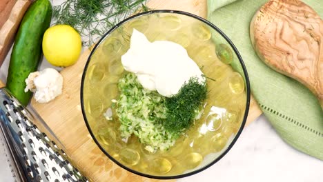 making tzatziki greek or mediterranean sauce or dip for a party. dip or sauce in a green bowl on a wooden cutting board with dill, green napkin and a grater. grinding black pepper into tzatziki sauce.