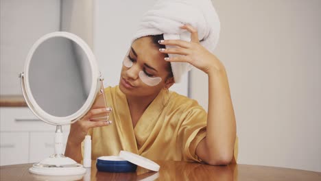 closeup portrait of beautiful woman after bath with towel on head puts patches under the eyes from wrinkles and dark circles. attractive female in bathrobe makes beauty treatments on the face at home.