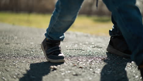 a close-up of an adult and a child wearing jeans and boots walking on a pavement, their shadows are reflected on the ground, and the pavement shows a minor pothole