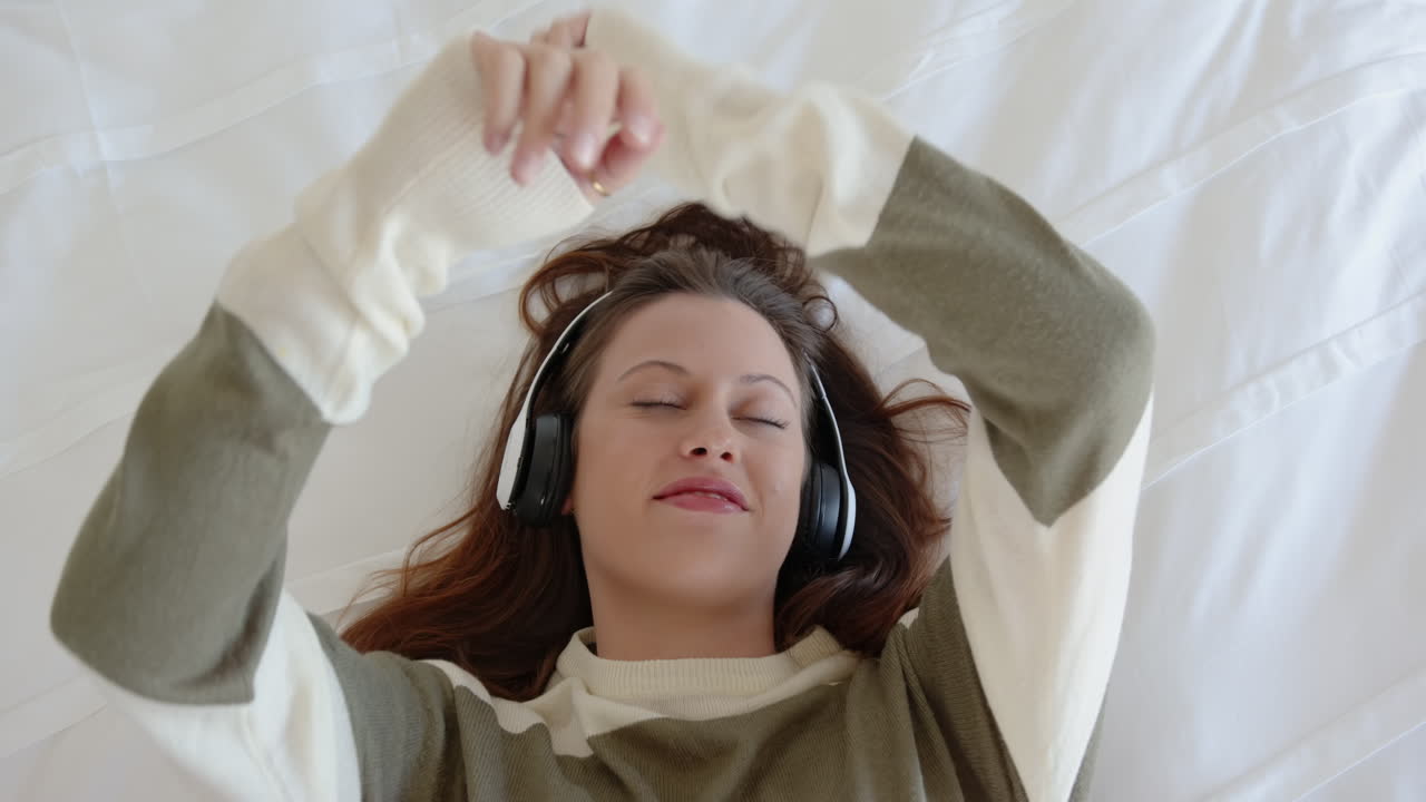 Relaxing on bed, young woman listening to music with headphones, eyes closed