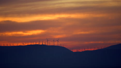 As-a-brilliant-sunset-lights-the-sky,-the-silhouette-of-hundreds-of-wind-turbines-generate-clean-energy---slow-motion