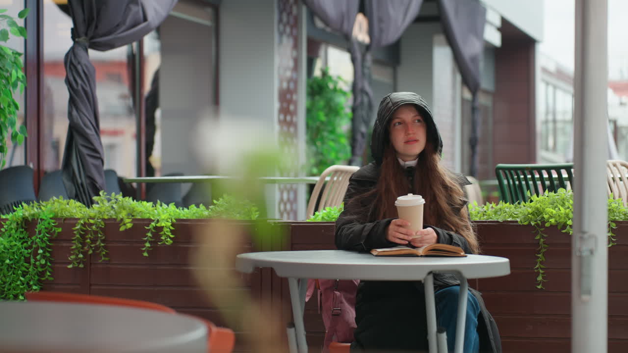 Donna incappucciata che sorseggia un latte macchiato in un bar, seduta a un tavolino circondato da piante pensili e vetrine di negozi urbani, leggera pioggerellina che crea un'atmosfera accogliente, ritratto spontaneo che cattura un momento di riflessione