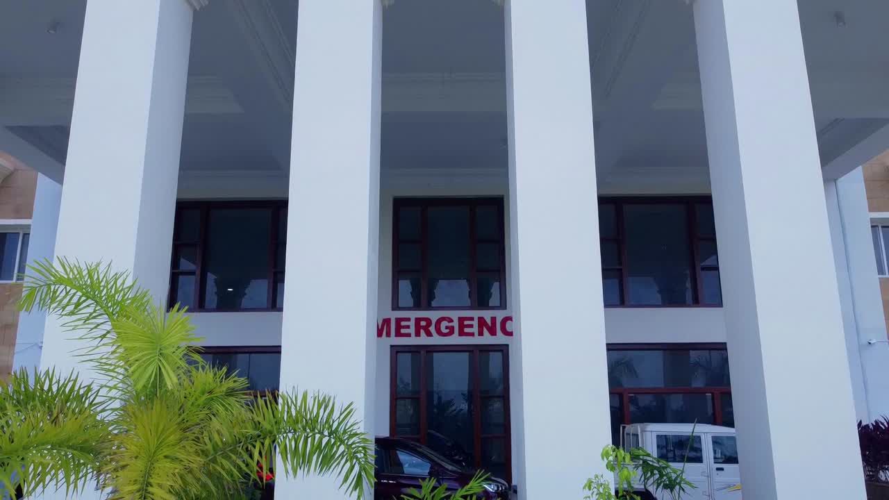 Uma tomada limpa com grua revelando a entrada de emergência de um grande e moderno hospital na Índia. A arquitetura imponente apresenta grandes colunas brancas, portas de vidro e uma ambulância estacionada nas proximidades.