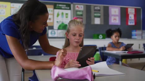 Diverse-smiling-female-teacher-helping-schoolgirl-sitting-in-classroom,-using-tablet