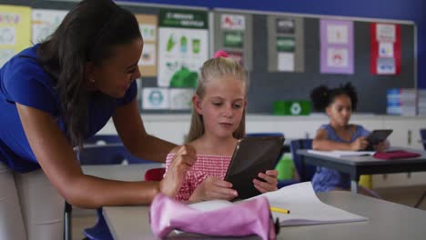 diverse smiling female teacher helping schoolgirl sitting in classroom, using tablet