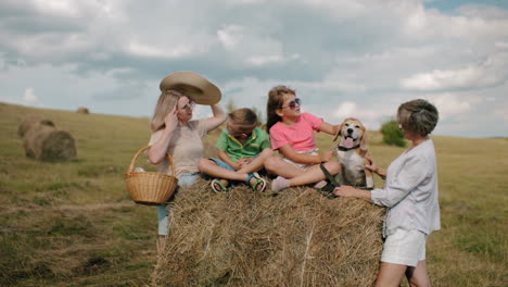 la familia disfruta de un picnic en el campo mientras la abuela frota la cabeza del perro, los niños se sientan en el pajar, la mujer lleva la canasta, el cielo de verano por encima, los campos verdes se extienden en la distancia
