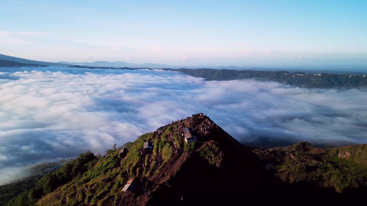 Pessoas observando o nascer do sol em cume de montanha vulcânica com vista panorâmica sobre cratera e mar de nuvens em luz dourada quente através do terreno montanhoso de Bali, filmagem aérea cinematográfica de drone.